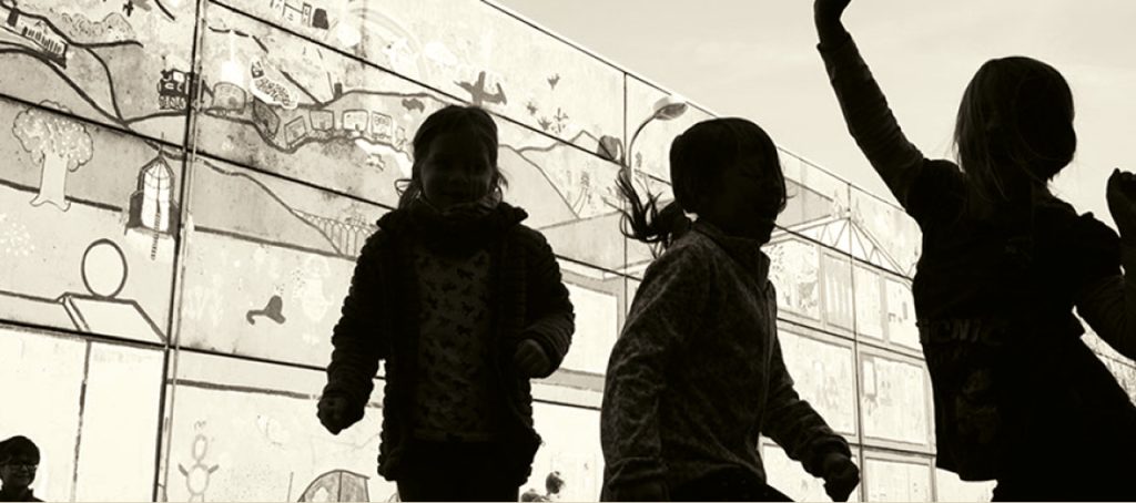Silhouetten von drei Kindern, die vor einer bemalten Wand entlanglaufen, ein Kind streckt den Arm hoch
