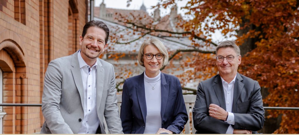Der Stiftungsvorstand der DEUBENER Stiftung, zwei Männer und eine Frau, lächelnd vor einem Backsteingebäude im Herbst